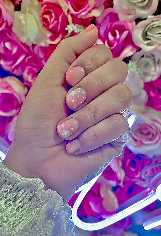 Uñas cortas rosas con glitter y detalles blancos frente a un muro de rosas y neón