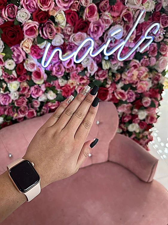 Rincón del estudio: muro de rosas rosas y rojas con letrero de neón blanco que dice nails y sillón de terciopelo rosa; una clienta muestra sus uñas negras